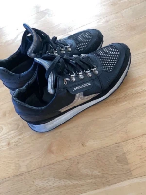 Svarta Dsquared2 skor - Snygga svarta sneakers från Dsquared2 med silverfärgade detaljer och metallhål för snörningen. Skorna har en vit sula och meshpartier framtill för extra ventilation. Perfekta för dig som vill ha en unik och trendig look.