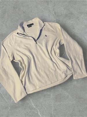 Vit halfzip från Polo Ralph Lauren - Snygg vit halfzip-tröja från Polo Ralph Lauren med blå logga broderad på bröstet. Tröjan har hög krage med dragkedja och är tillverkad i mjukt material som känns skönt mot huden. Perfekt för en clean och avslappnad stil.