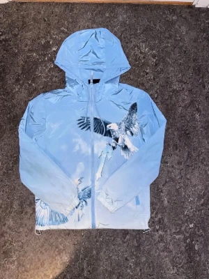 Trapstar ’ eaglewindbreaker - Cool blå vindjacka med huva och dragkedja, prydd med stora fågelmotiv på både fram- och baksidan. Jackan har ett tryck med texten 'TRAPSTAR' på ryggen och är tillverkad i ett lätt, vindavvisande material. Perfekt för dig som vill sticka ut med en unik design.