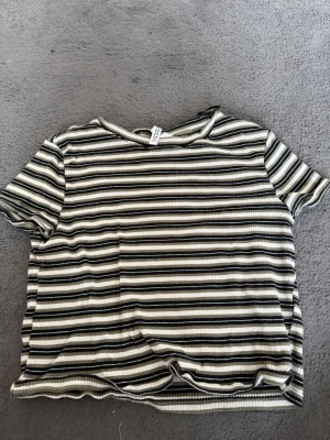 Randig ribbad topp från H&M - Säljer en ribbad topp från H&M med svart, vit och gråa horisontella ränder. T-shirten har korta ärmar och en enkel rund halsringning. Materialet är mjukt och stretchigt, perfekt för dig som gillar en chill och snygg stil.