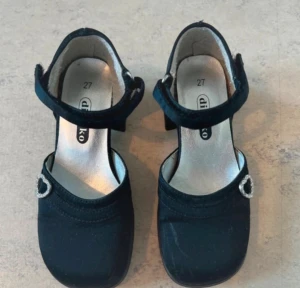 Svarta pumps med hjärtspänne D'Ko - Snygga svarta pumps från D'Ko med rund tå och bred klack. Skorna har ett dekorativt hjärtformat spänne på remmen och en klassisk siluett. Insidan är ljus med märkets logga tryckt på sulan. Perfekta för dig som gillar en söt och stilren look.