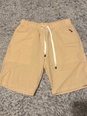 Linne, Beige shorts. Passar som small. - Snygga beige shorts med elastisk midja och vit snörning framtill. Shortsen har sidofickor och en bakficka, perfekta för sommardagar. Tillverkade i ett mjukt och lätt material som känns skönt mot huden.