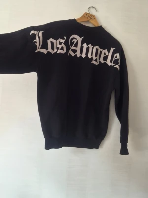 Svart oversized sweatshirt Los Angeles - Cool svart oversized sweatshirt från Black Squad i storlek XS. Stor vit text 'Los Angeles' på ryggen i gotisk stil. Tillverkad i mjukt material, perfekt för en avslappnad streetwear-look. Rund halsringning och ribbade muddar vid ärmar och nederkant.