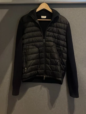 Svart pufferjacka från Moncler - Snygg svart pufferjacka från Moncler med quiltad front och stickade ärmar. Jackan har två dragkedjeförsedda fickor och hög krage. Perfekt för kalla dagar och ger en clean, modern look. Materialmix med dunfylld front och mjuka ärmar. Kvitto finns 