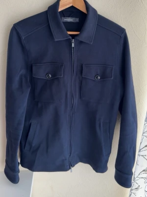 Mörkblå overshirt från dressman - Stilren mörkblå overshirt från Dressman med dragkedja framtill och två bröstfickor med knappar. Klassisk krage och långa ärmar med knappdetalj vid manschetten. Perfekt att bära som lager över t-shirt eller hoodie även perfekt för sommarkvällar!
