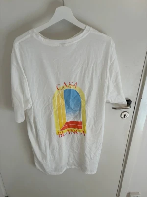 Vit Casa Blanca t-shirt med tryck - Vit t-shirt från Casa Blanca med färgglatt grafiskt tryck på ryggen i blått, gult och rött. Klassisk rund hals och korta ärmar. Perfekt för dig som gillar stilrena plagg med en twist. Materialet är mjuk bomull och passformen är normal.