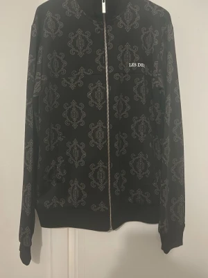 Les Deux fullzip - Snygg svart fullzip från Les Deux med hel dragkedja och unikt vitt mönster över hela plagget. Diskret Les Deux-logga på bröstet. Tillverkad i mjukt material och har ribbade muddar vid ärmslut och nederkant. Perfekt för en trendig och avslappnad stil.