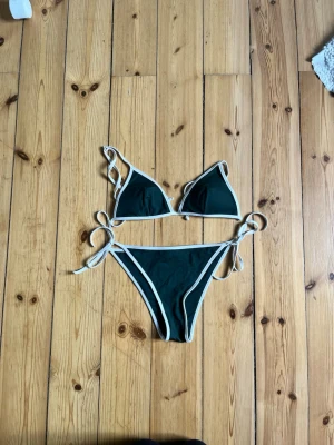 Mörkgrön bikini med vita kanter - Snygg mörkgrön bikini med vita kanter och knyt i sidorna samt axelbanden. Klassisk trekantsmodell på överdelen och matchande underdel. Perfekt för lata dagar på stranden eller poolen.