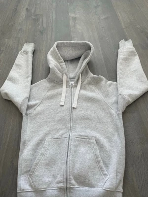 Ljusgrå zip-hoodie med fickor - En stilren ljusgrå hoodie med dragkedja framtill, stora kängurufickor och justerbar huva med vita snören. Tillverkad i mjukt sweatshirtmaterial och har ribbade muddar vid ärmslut och nederkant. Perfekt för en avslappnad look.
