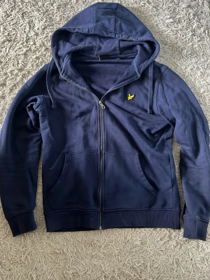 Marinblå hoodie från Lyle & Scott - Snygg marinblå hoodie från Lyle & Scott med dragkedja framtill och två stora fickor. Klassisk gul logga på bröstet och justerbar huva med snören. Tillverkad i mjuk bomullsblandning och har ribbade muddar vid ärmar och nederkant.