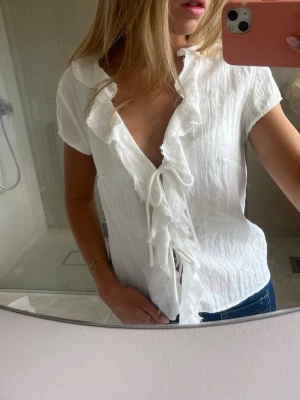 Weekday blus - Säljer en söt vit blus med volangkantad V-ringning och knyt framtill. Blusen har korta ärmar och är gjord i ett lätt, krinklat tyg som ger en avslappnad känsla. Perfekt att styla med jeans eller kjol för en trendig look.