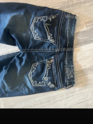 Mörkblå jeans med broderade fickor - Snygga mörkblå jeans från Truth be told Jeans med unika broderade detaljer på bakfickorna och kontrastsömmar. Klassisk femficksmodell och bootcut passform. Perfekta för dig som vill sticka ut lite extra med coola detaljer.
