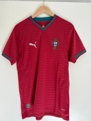 Portugal fotbollströja Puma röd - Snygg röd Portugal fotbollströja från Puma med gröna detaljer vid krage och ärmslut. Tröjan har korta ärmar, broderad Puma-logga och Portugals landslagsemblem på bröstet. Tillverkad i lätt och ventilerande material, perfekt för träning eller match.