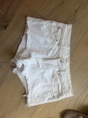 Vita fransiga jeansshorts från Gina 34 - Säljer ett par vita jeansshorts från gina tricot med råa, fransiga benslut och klassisk femficksdesign. Shortsen har knapp och dragkedja framtill samt en hög midja för en snygg siluett. Perfekta för varma dagar och enkel att matcha.