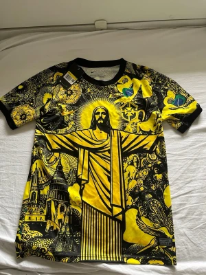  Brasilien fotbollströja  - Unik t-shirt med stort tryck av Kristusstatyn i Rio, detaljerade illustrationer av brasilianska symboler och byggnader. Färgstark design i gult och svart med inslag av grönt och blått. Rund hals, korta ärmar och tillverkad i mjuk polyester. Perfekt för dig som vill sticka ut. Storleken är S men passar även M