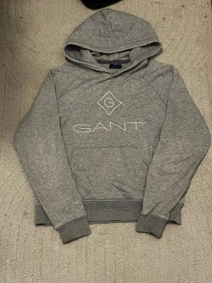 Grå hoodie från GANT med logga - Snygg gråmelerad hoodie från GANT med stor vit logga och emblem på bröstet. Klassisk huva och känguruficka framtill. Tillverkad i mjukt bomullsmaterial med ribbade muddar vid ärmslut och nederkant. Perfekt för en avslappnad och stilren look.