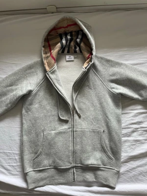  Burberry zip hoodie - Snygg grå zip hoodie från Burberry med klassiskt rutigt foder i huvan. Hoodien har två stora fickor framtill, dragsko i huvan och ribbade muddar vid ärmslut och nederkant. Perfekt för en chill och stilren look.