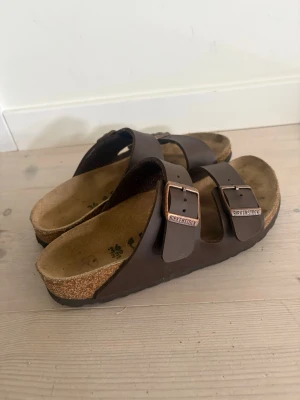 Bruna Birkenstock sandaler - Säljer ett par klassiska bruna Birkenstock sandaler med två breda remmar och justerbara metallspännen. Fotbädden är i kork och sulan är i gummi för extra komfort. Perfekta för en avslappnad och stilren look.