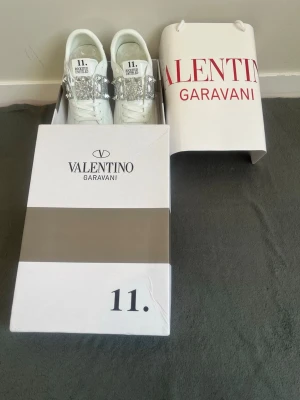Valentino Rockstud Untitled sneakers - Snygga vita sneakers från Valentino Garavani med silverfärgade nitar och glitterdetaljer över snörningen. Modellen heter Rockstud Untitled och har en clean, modern look med exklusiv känsla. Perfekta för dig som vill sticka ut med stilrena men ändå unika skor.