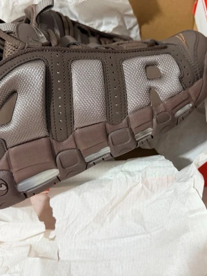 Nike Air More Uptempo bruna sneakers - Nike Air More Uptempo sneakers i en brun färg med stora AIR-bokstäver på sidan. Skorna har en chunky sula, mesh och syntetmaterial samt flera Swoosh-detaljer. Perfekta för dig som gillar retro och streetwear-vibe.