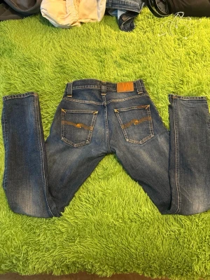 Nudie Jeans W31 & L32 - Säljer ett par klassiska blå jeans från Nudie Jeans med raka ben och snygga slitningar. Jeansen har fem fickor, orange sömmar och Nudie-logga på bakfickan. Tillverkade i bomull med en skön passform och tidlös tvätt.