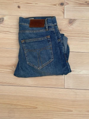 Blå raka jeans från Nudie Jeans - Säljer ett par klassiska blå jeans från Nudie Jeans med raka ben och fem fickor. Jeansen har en snygg tvätt och detaljerade sömmar i kontrastfärg. Tillverkade i slitstark denim med läderpatch bak i midjan. Perfekta för en avslappnad och stilren look.