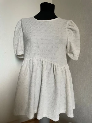 Vit kort klänning med puffärm - Supersöt vit kort klänning med puffärmar och rund hals. Klänningen har en strukturerad yta och ett lätt utställt kjolparti som ger en avslappnad men ändå chic look. Perfekt för sommarens alla tillfällen!