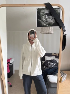 Vit hoodie med dragkedja och huva - Säljer en enkel och clean vit hoodie med dragkedja framtill och stor ficka på magen. Hoodien har justerbar huva med snören och ribbade muddar vid ärmar och nederkant. Perfekt för chill dagar eller när du vill ha en basic look.