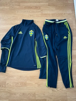 Svenskt träningsset Adidas blå/gul - Säljer ett blått träningsset från Adidas med Sveriges landslagsemblem. Setet består av en långärmad tröja med hög krage och matchande byxor, båda med gula detaljer och Adidas-logga. Perfekt för fotbollsträning eller chill. Materialet är mjukt och stretchigt.