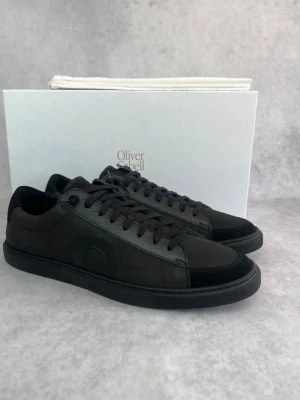 Svarta sneakers från Oliver Cabell - Stilrena helsvarta sneakers från Oliver Cabell med lågt skaft. Skorna har ovandel i mocka och läder, ton-i-ton snörning och diskret cirkeldetalj på sidan. Perfekta för dig som gillar minimalistisk och clean stil.