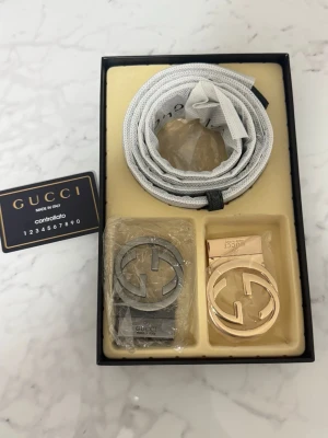 GG bälte med två spännen - Säljer ett GG bälte i ljusgrått tyg med två utbytbara GG-spännen, ett i silver och ett i guld. Bältet har klassisk design och levereras i originalförpackning med kontrollkort. Perfekt accessoar för att lyfta din outfit.  Inte använt helt ny
