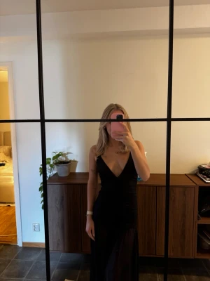 Svart långklänning med smala axelband - Elegant svart långklänning med djup v-ringning och tunna axelband. Klänningen har en figurnära passform och öppen rygg, vilket ger en stilren och modern look. Klänningen är i nyskick med prislappen kvar