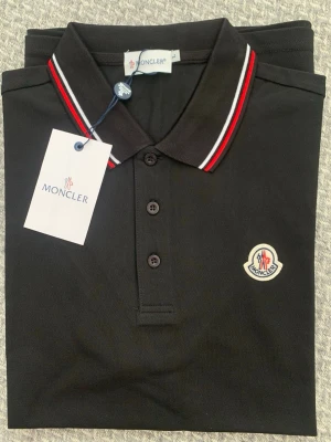 Svart Moncler pikétröja med ränder - Snygg svart pikétröja från Moncler med klassisk krage och knäppning framtill. Kragen har röda och vita ränder för en sportig touch och bröstet pryds av Monclers ikoniska logotyp. Perfekt till jeans eller shorts för en clean look.
