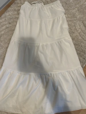 Vit långkjol med volanger och knyt - Säljer en vit långkjol med volangdetaljer och knyt i midjan. Kjolen har en luftig och flowy passform, perfekt för varma dagar. Tillverkad i ett mjukt tyg som ger en skön känsla. Enkel att styla med både toppar och sneakers.