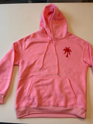 Rosa hoodie Palm Puff med tryck - Cool rosa hoodie från Palm Puff med röd palmtryck på bröstet och stort surf-inspirerat motiv på ryggen. Hoodien har huva med dragsko och en stor magficka. Materialet är mjukt och bekvämt, perfekt för chill dagar. Snygga ribbade muddar på ärmar och nederkant.