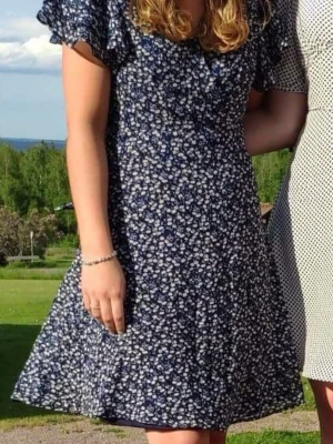 Blommig blå klänning från Gina Tricot - Mörkblå klänning med vita småblommiga mönster från Gina Tricot. Klänningen har korta volangärmar, v-ringning och lätt utställd kjol. Perfekt för sommardagar och har en feminin, avslappnad stil. Materialet känns lätt och luftigt.