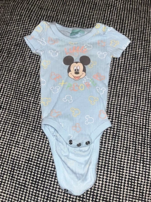 Blå Disney body med Musse Pigg - Söt ljusblå body från Disney med Musse Pigg-tryck framtill och texten 'Little'. Kortärmad modell med färgglada Musse Pigg-huvuden över hela plagget och tryckknappar i grenen. Perfekt för dig som gillar gulliga och lekfulla motiv.