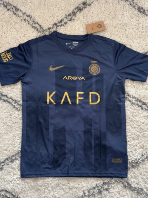 Blå Al Nassr Ronaldo matchtröja Nike - Snygg mörkblå Al Nassr matchtröja från Nike med guldiga detaljer och tryck. Framsidan har klubbmärke, Nike-logga och sponsortryck i guld. Baksidan har namnet 'Ronaldo' och nummer 7 i guld. Tillverkad i lätt och ventilerande material, perfekt för fotboll eller träning.