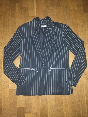 Svart kavaj med vita ränder Noisy May - Snygg svart kavaj från Noisy May med vita vertikala ränder och två dragkedjefickor framtill. Klassisk krage och figurnära passform. Perfekt för en trendig och stilren look. Materialet känns mjukt och följsamt.