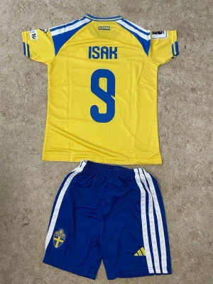 Sverige matchställ Isak #9 Adidas - Säljer ett officiellt Sverige matchställ från Adidas med Isak och nummer 9 på ryggen. Tröjan är gul med blå detaljer och korta ärmar, byxorna är blå med vita ränder och Sveriges landslagsemblem. Perfekt för fotbollsträning eller match.