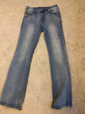 Ljusblå bootcut jeans med slitning - Säljer ett par ljusblå bootcut jeans med klassisk femficksdesign och en liten slitning på ena benet. Jeansen har en något utsvängd passform nedtill och snygga tvättade detaljer. Perfekta för en avslappnad och trendig look.