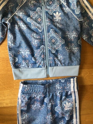 set Adidas - Säljer ett blått loungewear-set från Adidas med unikt orientaliskt mönster i vitt och ljusrosa. Setet består av ziptröja och byxor med vita ränder längs ärmar och ben samt Adidas-logga på både tröja och byxa. Perfekt för chill dagar 