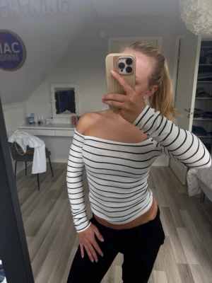 Randig offshoulder topp i ribbad bomull - Säljer en vit ribbad offshoulder topp med svarta horisontella ränder. Toppen har lång ärm och sitter tight mot kroppen för en snygg siluett. Perfekt för dig som gillar en enkel men trendig stil.