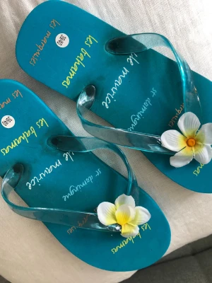 Turkosa flip-flops med blomma - Säljer ett par turkosa flip-flops från Les Marquises med transparenta remmar och dekorativa vita blommor med gul mitt. Perfekta för stranden eller sommardagar. Tillverkade i syntetmaterial och har en färgglad, tropisk vibe. Se bilder.