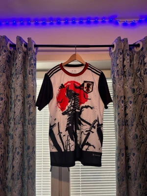 Japan Adidas fotbollströja samurai - Snygg Japan fotbollströja från Adidas med svartvita färger, svart samurai-tryck och röd sol på bröstet. Tröjan har svarta ärmar, vita axelränder och Japan-emblem på vänster sida. Tillverkad i lätt och ventilerande material, perfekt för tränings eller match.