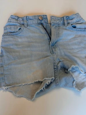 Ljusblå jeansshorts med fransig kant - Säljer ett par ljusblå jeansshorts med fransig, rå kant och klassisk femficksdesign. Shortsen har dragkedja och knapp framtill samt slitna detaljer för en avslappnad look. Perfekta för varma dagar och enkel att matcha med allt.