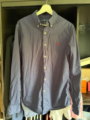 Mörkblå skjorta från Ralph Lauren - Klassisk blå skjorta från Ralph Lauren i slim fit med knappkrage och broderad röd logga på bröstet. Tillverkad i lätt bomullstwill som känns luftig och skön. Långärmad modell med knappar framtill och vid ärmslut. Skönt material för sommaren