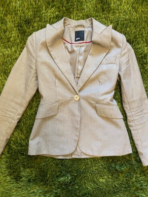 Beige figursydd tweed kavaj från Gina Tricot - Stilren beige figursydd tweed kavaj från Gina Tricot med klassisk passform, ett guldigt knappslå och två fickor framtill. Kavajen har smal krage och är helfodrad med slätt tyg.