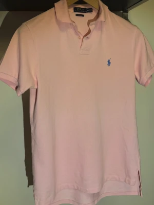 Ljusrosa pikétröja från Ralph Lauren - Klassisk ljusrosa pikétröja från Ralph Lauren med blå logga broderad på bröstet. Tröjan har korta ärmar, krage och knappar vid halsen. Tillverkad i mjuk bomull som känns skön mot huden.