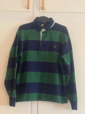 Randig rugbytröja från Gant - Klassisk långärmad rugbytröja från Gant med breda marinblå och gröna ränder. Skjortan har vit krage, knappar i halsen och en liten broderad logga på bröstet. Tillverkad i mjuk bomull för en bekväm och avslappnad stil.